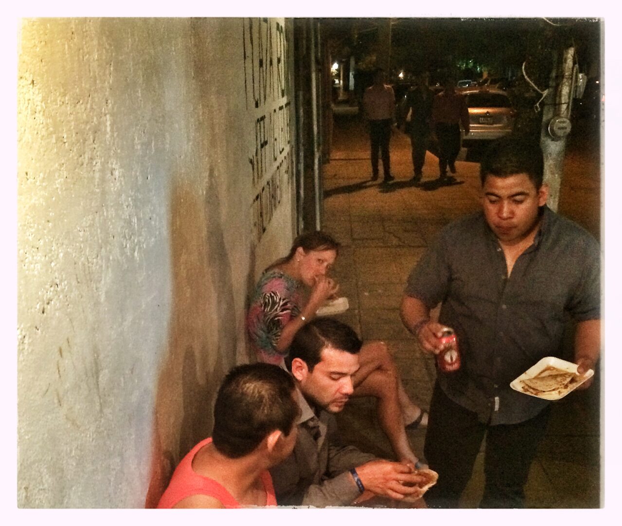 A group of people sitting on a sidewalk eating food.