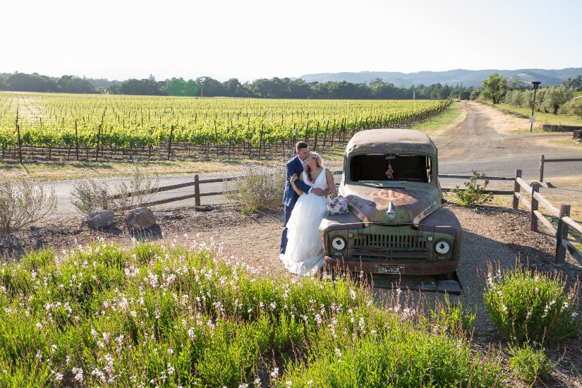 A bride and groom pose next to an old truck in a vineyard.