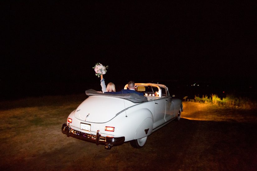 A bride and groom riding in a vintage car at night.