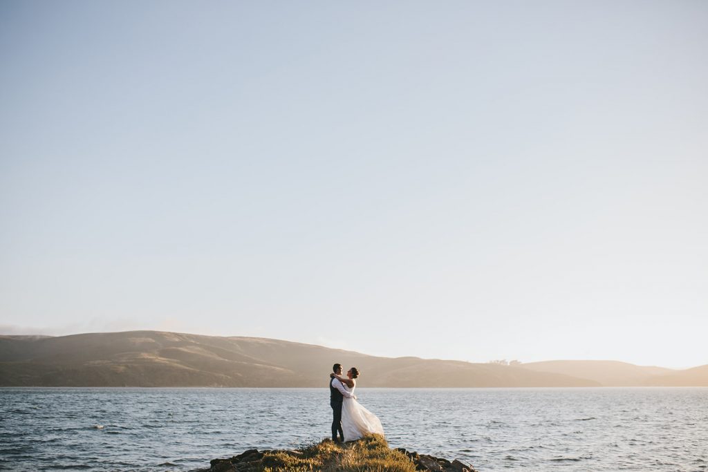 A bride and groom standing on a rock overlooking the ocean.