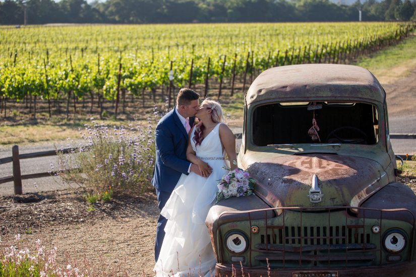 A bride and groom pose next to an old truck in a vineyard.