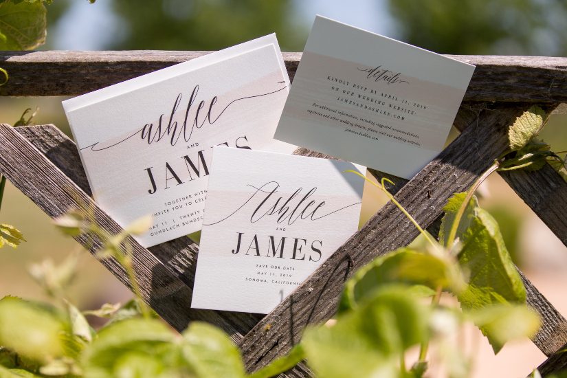 Three wedding invitations hanging on a wooden fence.