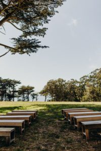 An outdoor wedding ceremony with wooden benches and a tree in the background.