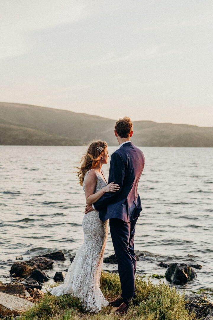 A bride and groom standing by the water at sunset.