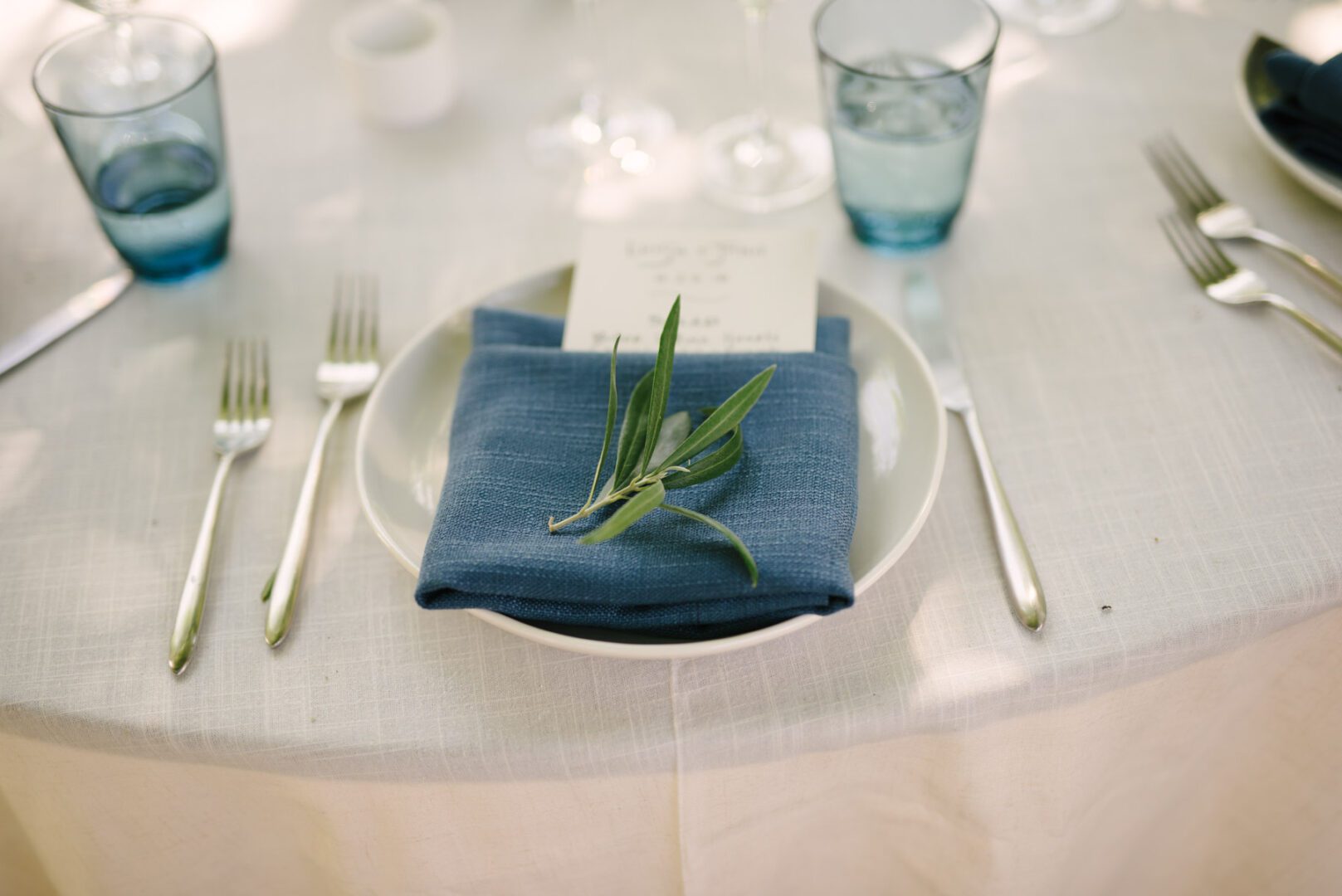 A table setting with a blue napkin and a sprig of olives.