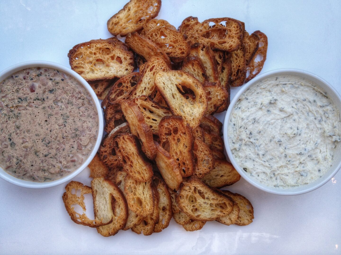 Two bowls of dip with bread and crackers on a white plate.