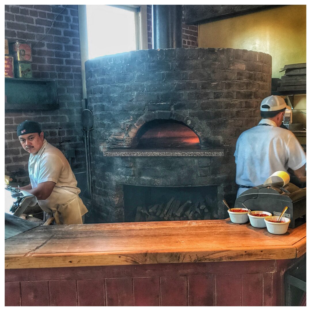Two men standing in front of a pizza oven.