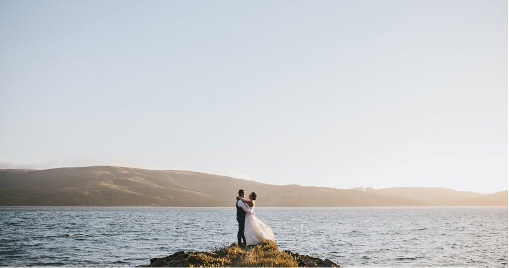A bride and groom standing on top of a rock in front of the ocean.