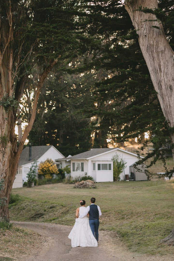 A bride and groom walking down a dirt road in front of a large tree.