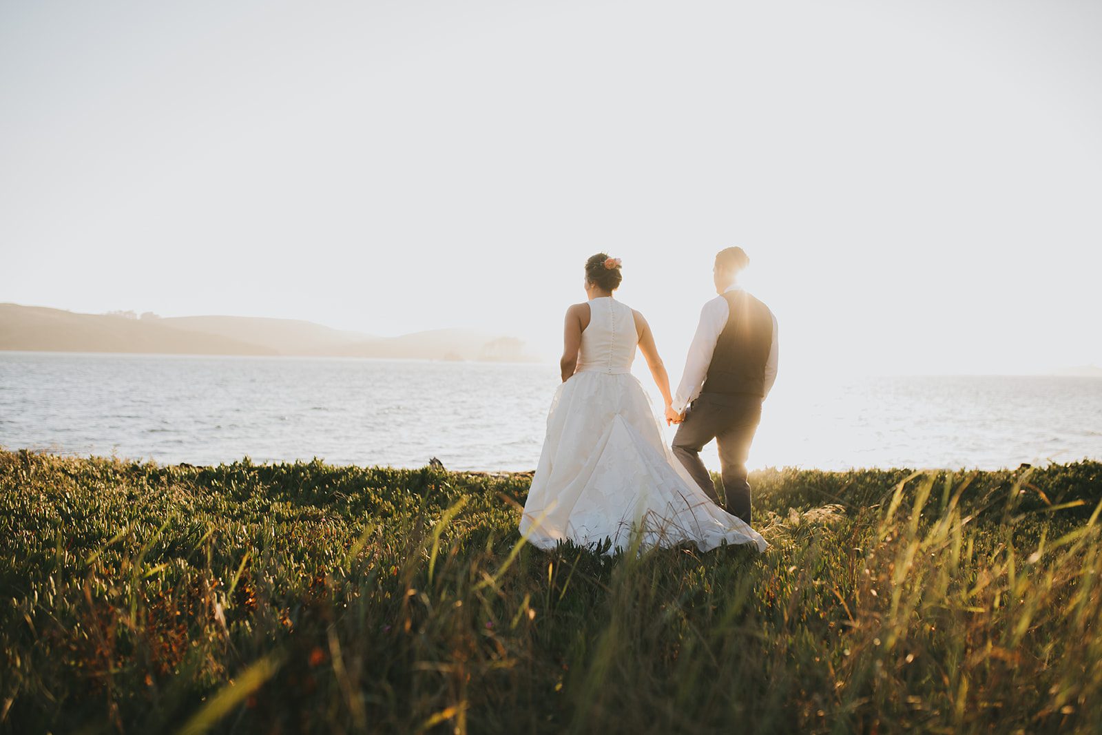 A bride and groom standing in a grassy field near the water.