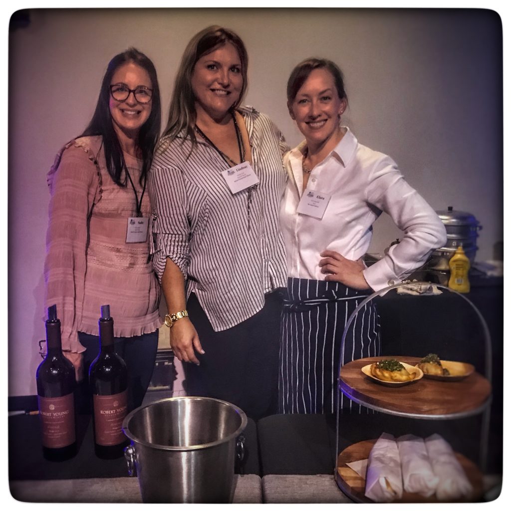 Three women standing in front of a table with wine and food.