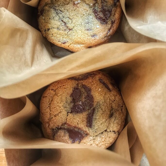 Two chocolate chip cookies in brown paper bags on a table.