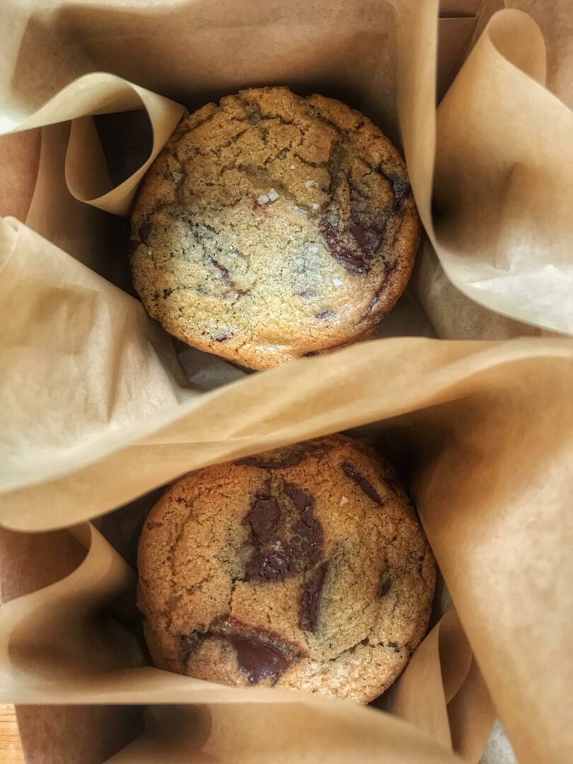 Two chocolate chip cookies in brown paper bags on a table.