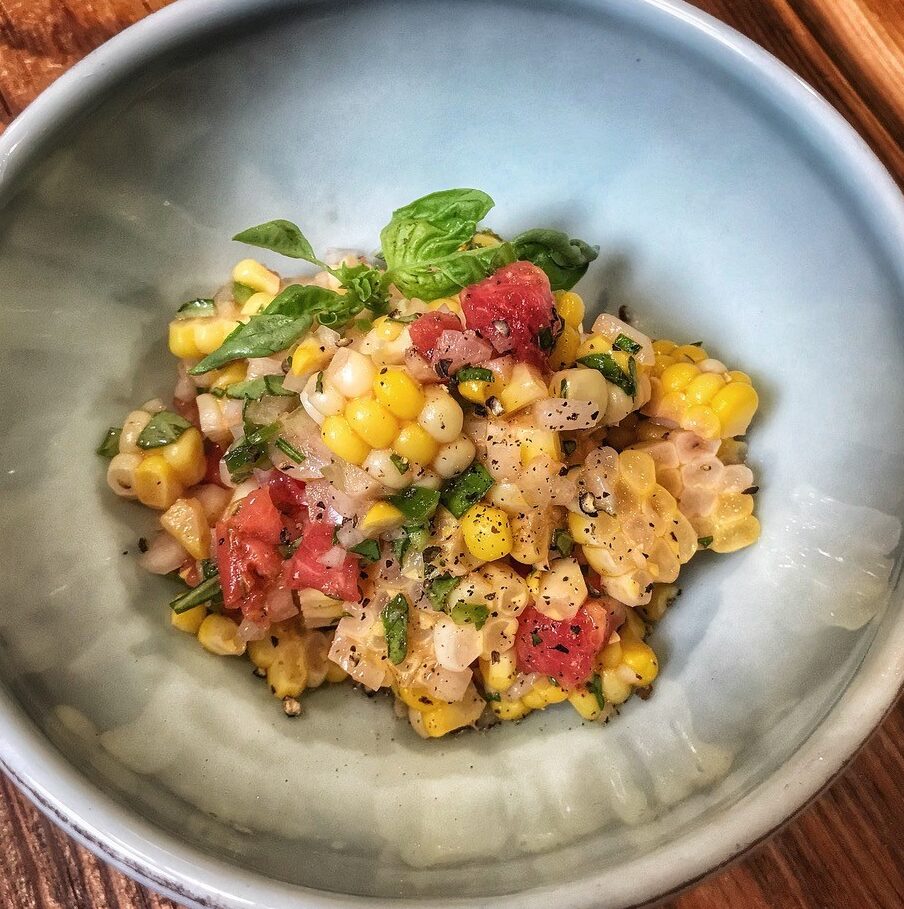 A bowl of corn, tomatoes and basil on a wooden table.